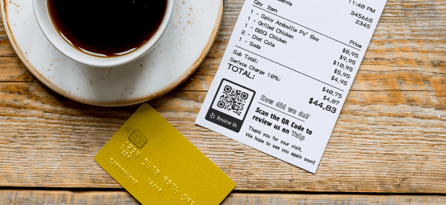 A cup of coffee, a credit card, and a receipt with a QR Code for Yelp reviews on a wooden table.