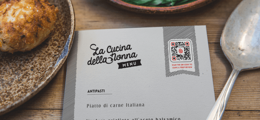 A plate, spoon, and menu with a Yelp QR Code on a wooden table.